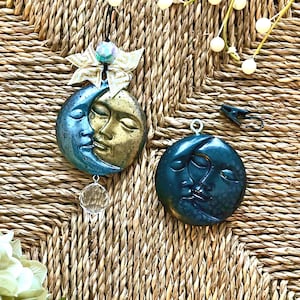 Könnte beinhalten: Zwei mondförmige Ornamente. Eines ist blau und goldfarben mit einem Gesicht, einem Kristall und einer dekorativen Schleife. Das andere ist dunkelblau mit einem Gesicht. Sie hängen vor einem gewebten Hintergrund.