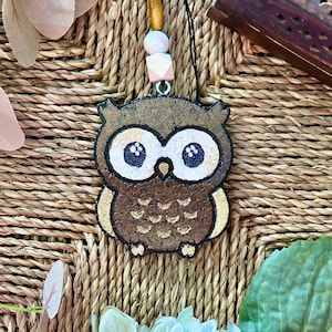 Op de afbeelding: Een bruine uil ornament met gouden accenten en witte ogen. De uil is gemaakt van kurk en heeft een rustieke uitstraling.