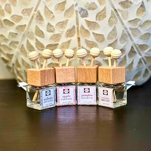 Op de afbeelding: Vier kleine glazen flesjes met houten doppen en decoratieve houten balaccenten. Elk flesje heeft een label met een andere geurnaam: Velvet Orchard, Apple Caramel, Pumpkin Hearth en Crème Brûlée. De flesjes zijn in een rij gerangschikt.