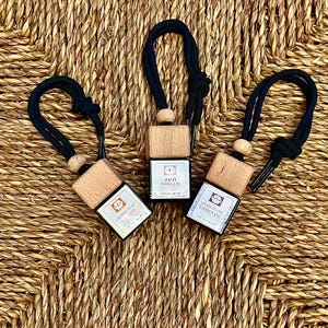 May include: Three wooden aromatherapy car air fresheners with black cords and small wooden beads. Each has a small, square label with text. The background is a woven, natural-colored material.