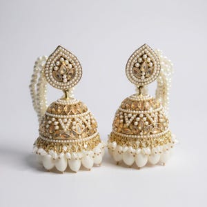 May include: A pair of gold-toned, ornate earrings with a teardrop-shaped top and a bell-shaped bottom. The earrings are embellished with small pearls and clear, faceted stones. A strand of pearls is attached to each earring.