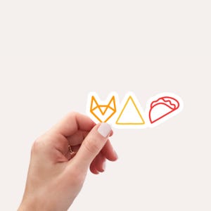 May include: A hand holding three colorful stickers. The stickers feature a geometric fox head, a triangle, and a taco. The fox and taco are outlined in orange and red, respectively, while the triangle is outlined in yellow.