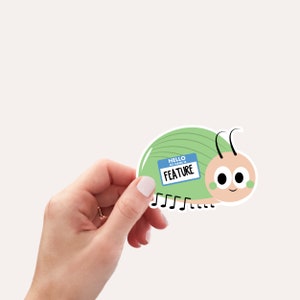 May include: A sticker featuring a cartoon caterpillar with a light green body and a light pink head. The caterpillar has black antennae and a name tag that reads "FEATURE". The sticker is held by a hand.