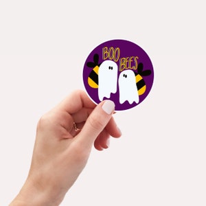 May include: A round sticker with a purple background features two cartoon ghosts with the text "BOO BEES" in yellow. The ghosts are positioned next to bee-like designs with black and yellow stripes. The sticker is held by a hand.