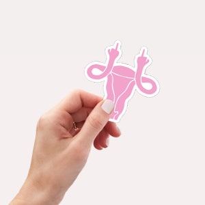 May include: Pink anatomical illustration of a uterus with two arms raised in a middle finger gesture. The sticker is held in a hand.