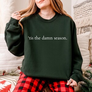 May include: Dark green sweatshirt with the phrase "'tis the damn season" in white lettering. The sweatshirt is a crew neck style. The person in the image is wearing red and black plaid pajama pants.