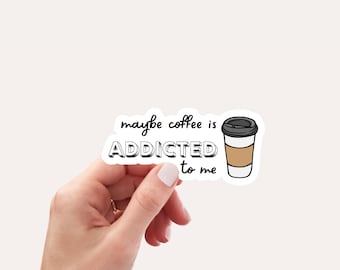 May include: A white sticker with the text "maybe coffee is ADDICTED to me" in black, outlined in white. A cartoon coffee cup is to the right. The sticker is held by a hand with a gold ring.