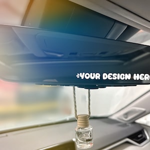 May include: A rear view mirror with a white sticker that says "YOUR DESIGN HERE" and a small, clear glass bottle hanging from it with a wooden cap.
