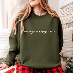May include: Olive green sweatshirt with the phrase "in my merry era" in white script. The sweatshirt is paired with red and black plaid pajama pants. The overall theme is festive and cozy.