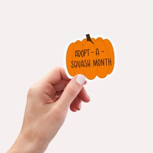 May include: An orange pumpkin-shaped sticker with the text "ADOPT-A-SQUASH MONTH" in black. The sticker is held by a hand with a gold ring and light pink nail polish.