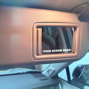 May include: A beige car sun visor with a rectangular mirror. The mirror has a white sticker that says "YOUR DESIGN HERE".