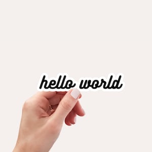 May include: A white sticker with black text that reads "hello world" being held by a hand.