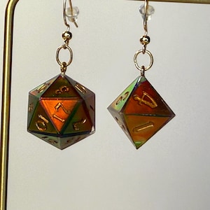 Peut inclure: Une paire de boucles d'oreilles pendantes composées de dés polyédriques aux reflets irisés changeants dans des tons orange, vert et doré. Les dés présentent des chiffres gravés de couleur dorée et sont suspendus à des crochets en métal doré.