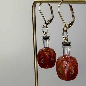 May include: A pair of dangle earrings featuring miniature potion bottle charms. Each charm is a translucent orange, multi-faceted polyhedral shape resembling a gaming die with gold numbers, topped with a clear neck and a black stopper on gold hooks.