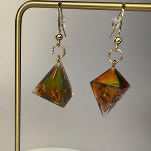 May include: A pair of drop earrings featuring translucent, multi-coloured iridescent polyhedral dice charms with gold-coloured numbers. The dice are suspended from gold-toned metal fishhook wires with small clear plastic backings for secure wear.