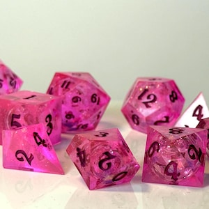 May include: A collection of pink and clear resin dice with black numbering. The dice are in various polyhedral shapes, including D4, D6, D8, D10, D12, and D20. The dice are scattered on a white surface.