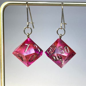 May include: A pair of D20 dice earrings. The dice are a vibrant pink with gold-coloured numbers. The earrings hang from gold-coloured ear wires. A unique accessory for tabletop gaming fans. The dice are approximately 2.5 cm in size.