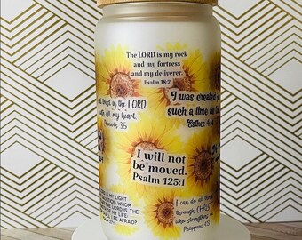 Puede incluir: Un vaso blanco con tapa de bambú y una pajita. El vaso tiene un diseño de girasoles y versículos bíblicos inspiradores, incluyendo "El SEÑOR es mi roca, mi fortaleza y mi libertador. Salmo 18:2", "Confío en el SEÑOR con todo mi corazón. Proverbios 3:5", "Fui creado para un tiempo como este. Ester 4:14", "No seré movido. Salmo 125:1", y "Todo lo puedo en Cristo que me fortalece. Filipenses 4:13".