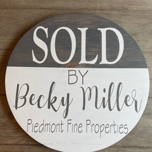 May include: A round wooden sign with a gray and white paint finish. The top half of the sign is gray with white text that reads "SOLD". The bottom half of the sign is white with gray text that reads "BY Becky Miller Piedmont Fine Properties".