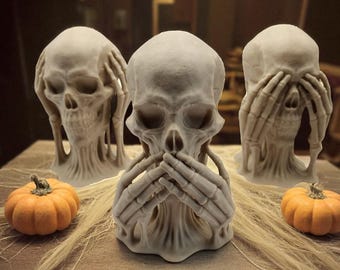 May include: Three off-white skull sculptures, each with hands covering the eyes, ears, and mouth, arranged on a table with two small orange pumpkins and decorative strands. The skulls are a Halloween or gothic theme.
