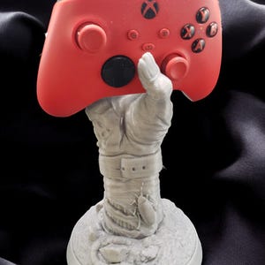 May include: A 3D printed stand in the shape of a hand holding a red Xbox controller. The hand is wrapped in bandages and has a belt around its waist. The base of the stand is a circular platform with a textured surface.