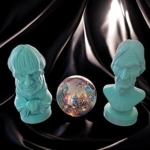 May include: Three teal blue ceramic busts of people with a sparkly iridescent glass ball in the center. The busts are on a black fabric background.