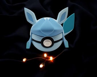 May include: A blue and white Pokemon figure of a blue Eevee with a Pokeball on its head. The figure is sitting on a black surface with a string of lights in front of it.