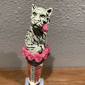 May include: A white tiger figurine with black stripes, wearing a pink flower crown, is a beer tap handle. The handle is decorated with a pink and orange design and the text "Easy Tiger".