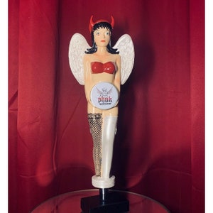 May include: A white ceramic beer tap handle with a red and white angel figure with wings and devil horns. The angel is holding a round white disc with the text "phuk" in red and black.