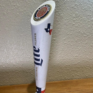 May include: White wooden beer tap handle with a blue and red Texas flag design. The top of the handle has a gold and red circular logo that reads "A Fine Pilsner Beer". The handle also has the text "The Original Miller" and "Pilsner" printed on it.
