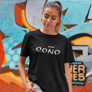 May include: Black t-shirt with the word "COÑO" in white script. The shirt is being worn in front of a colorful graffiti background. The t-shirt is a casual wear item.
