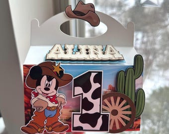 Puede incluir: Una caja de regalo blanca con temática de vaquero. La caja presenta un Mickey Mouse de dibujos animados con un atuendo de vaquero, un número uno con un patrón de piel de vaca, una rueda de carro de madera y un cactus. El nombre "ALINA" se muestra en la parte superior.