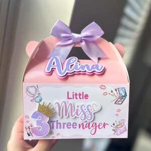 May include: A pink gift box with a purple bow and the name "Alina". The box is decorated with makeup-themed illustrations and the text "Little Miss Threenager" with a gold crown and the number 3.