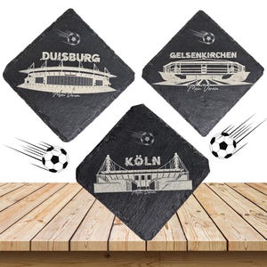 Könnte beinhalten: Drei schwarze Schieferuntersetzer mit weißen, geätzten Designs von Fußballstadien und Städtenamen: Duisburg, Gelsenkirchen und Köln. Jeder Untersetzer zeigt einen Fußball und die Worte "Mein Verein."