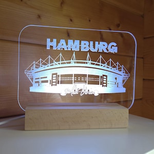 Könnte beinhalten: Eine rechteckige LED-Lampe aus klarem Acryl mit einer gravierten Stadion-Illustration und dem Schriftzug HAMBURG mit kleinen Figuren in den Buchstaben sowie dem Text Mein Verein darunter, montiert auf einem hellen Holzsockel.