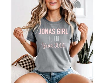 May include: Gray t-shirt with the text "JONAS GIRL TIL THE Year 3000" in pink lettering. The shirt has short sleeves and a crew neck. The model is wearing denim shorts.