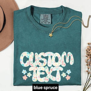 May include: A blue spruce t-shirt with the words "CUSTOM TEXT" in a colorful heart design. The shirt has a crew neck and short sleeves. A gold necklace with a star pendant is on the shirt. The shirt is size XL.