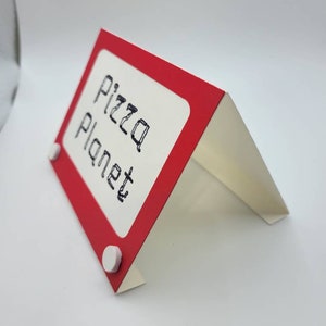 May include: Red and white cardboard sign with the text "Pizza Planet" written in black marker. The sign is shaped like a miniature Etch A Sketch.