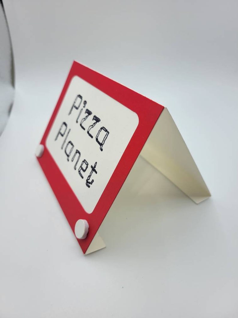 May include: Red and white cardboard sign with the text "Pizza Planet" written in black marker. The sign is shaped like a miniature Etch A Sketch.