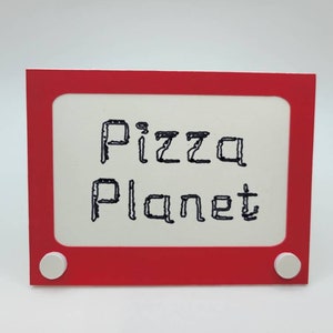 May include: A red plastic frame with white knobs, resembling a vintage Etch A Sketch toy. The frame has a white background with the words "Pizza Planet" written in black.
