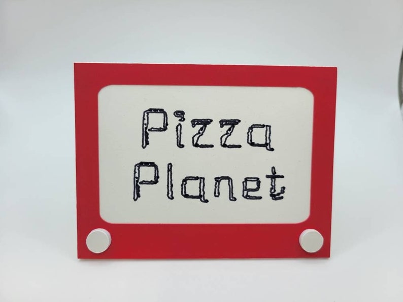 May include: Red plastic frame with white knobs, resembling a vintage Etch A Sketch toy. The frame has a white background with the words "Pizza Planet" written in black.