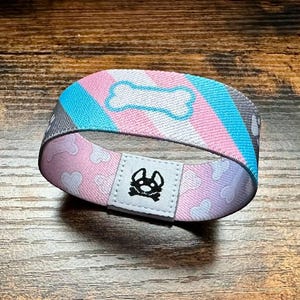 Puede incluir: Una pulsera con un patrón a rayas rosa, azul y gris. La pulsera tiene un diseño en forma de hueso blanco y un pequeño logotipo de huella de pata negra.