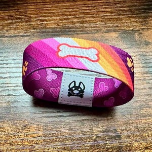 May include: A pink, orange, and white striped wristband with a white bone and paw print design. The wristband has a black dog silhouette on a white background.