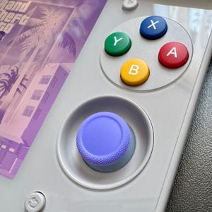 May include: A handheld gaming console with a light grey casing featuring a purple textured joystick and four coloured action buttons labelled X, Y, A, and B. The screen displays a purple-toned graphic with palm trees and the text Grand Theft Auto.