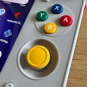 May include: A handheld gaming console with a light gray casing, featuring a bright yellow textured analog joystick and four primary action buttons labeled X, Y, A, and B in blue, green, red, and yellow colors respectively, alongside a digital screen.