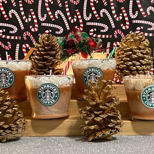 May include: Four brown and white Starbucks coffee scented candles in glass jars with a white lid. The candles are decorated with a white swirl and a green Starbucks logo. The candles are sitting on a wooden surface with pine cones.