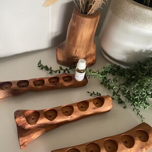 May include: Three elongated, natural-finish wooden holders, each featuring five circular indentations for essential oil bottles. The wood displays a rich, warm brown grain with organic, irregular edges. A small glass bottle labelled Balance sits in one holder.