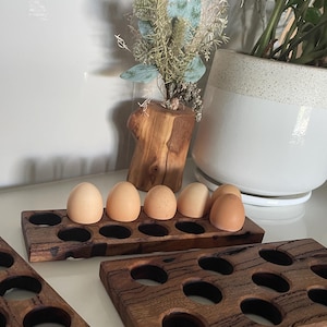 May include: Three wooden egg holders in different sizes. The largest holder has 20 round holes, the medium holder has 10 round holes, and the smallest holder has 8 round holes. The holders are made of dark brown wood with a natural wood grain. Six brown eggs are sitting in the medium-sized holder.