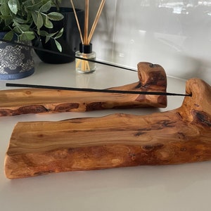 Two handcarved wooden incense holders, with black incense sticks visible. The wood is grainy with natural curves. Plants and an oil diffuser in the background of the image.