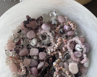 May include: A large white bowl filled with a collection of polished pink and clear stones, including several bracelets made from small, irregular stone chips and smooth, rounded pebbles in various shades of rose, white, and gray.